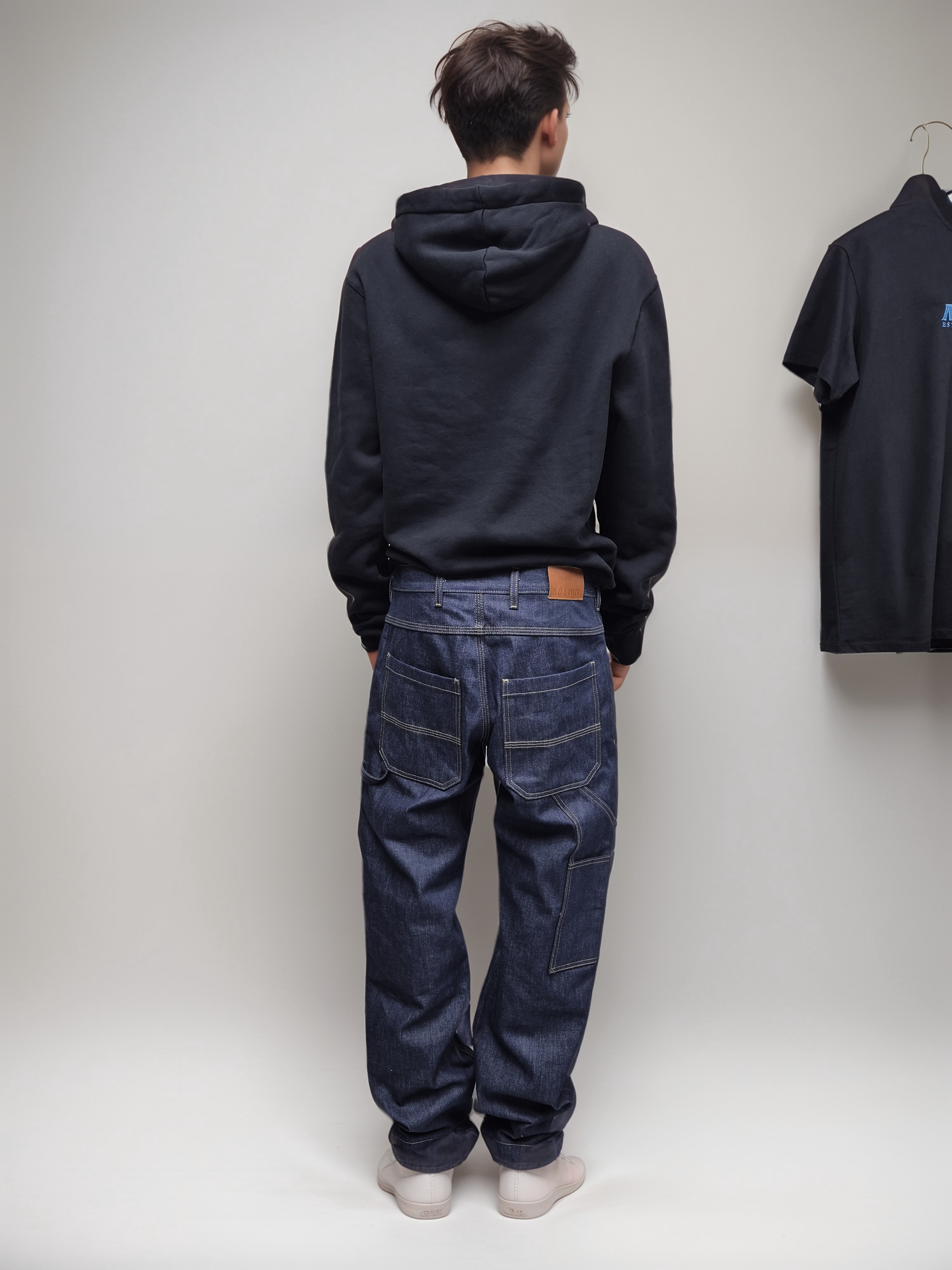 A person standing with their back to the camera wearing indigo blue boot cut leg carpenter workwear jeans and a dark hoodie.