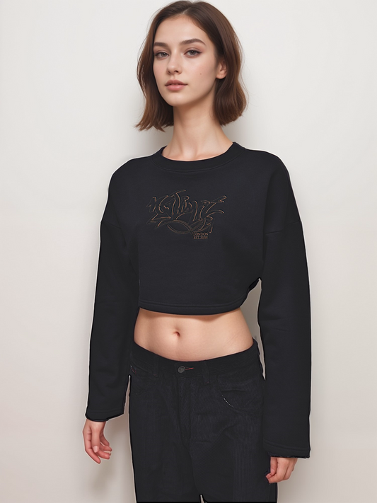 A woman wearing a black cropped pullover sweatshirt with gold embroidery, paired with black shorts.